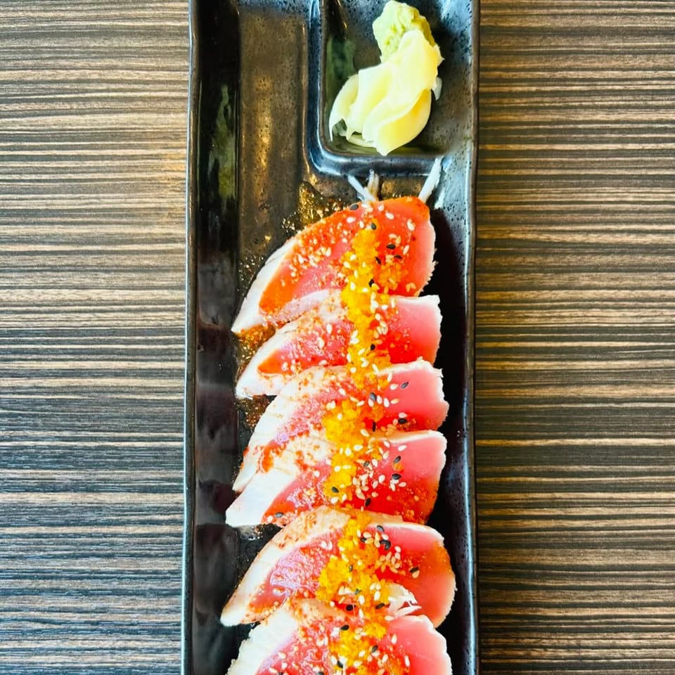 Seared tuna sashimi with orange roe, sesame seeds, and pickled ginger on a black rectangular plate.