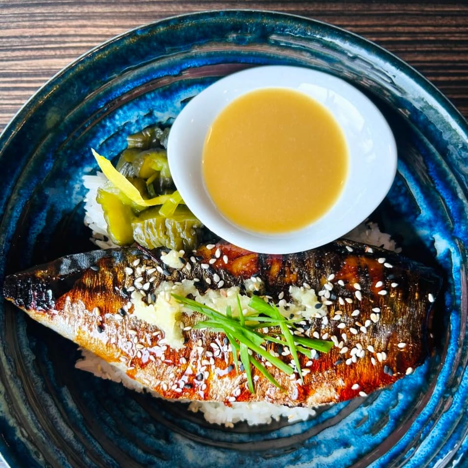 Grilled fish with sesame seeds, rice, pickled vegetables, and sauce in a blue bowl.