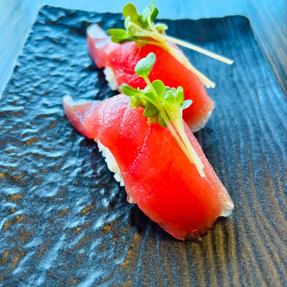 Two pieces of vibrant red tuna nigiri sushi, garnished with fresh green sprouts on a dark textured plate.